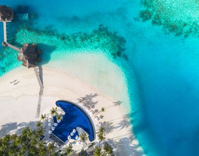 Aerial view of a tropical paradise with turquoise waters, white sandy beach, a curving pool, umbrellas, and a thatched-roof gazebo on a pier.