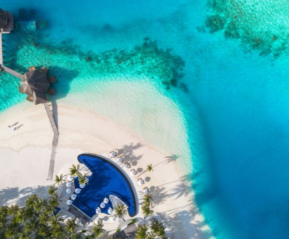Aerial view of a tropical paradise with turquoise waters, white sandy beach, a curving pool, umbrellas, and a thatched-roof gazebo on a pier.