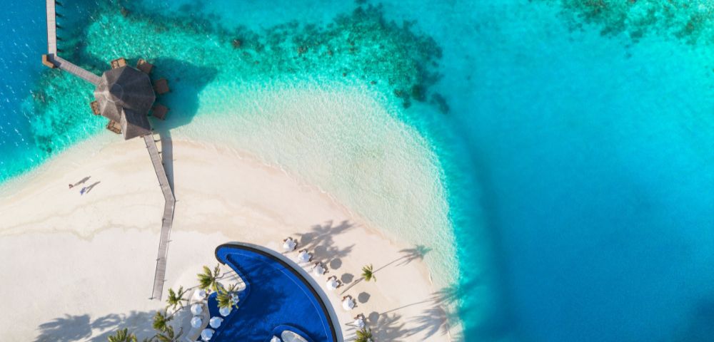 Aerial view of a tropical paradise with turquoise waters, white sandy beach, a curving pool, umbrellas, and a thatched-roof gazebo on a pier.