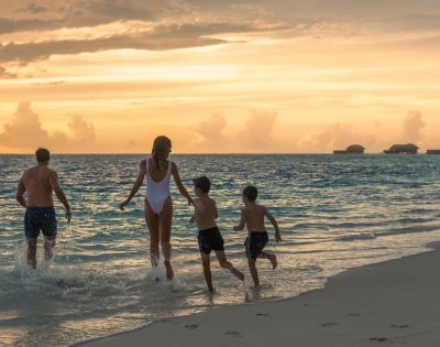 A family of four, including two children, joyfully runs into the ocean at sunset. The sky glows orange, and overwater bungalows are visible in the distance.