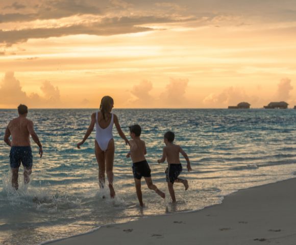 A family of four, including two children, joyfully runs into the ocean at sunset. The sky glows orange, and overwater bungalows are visible in the distance.