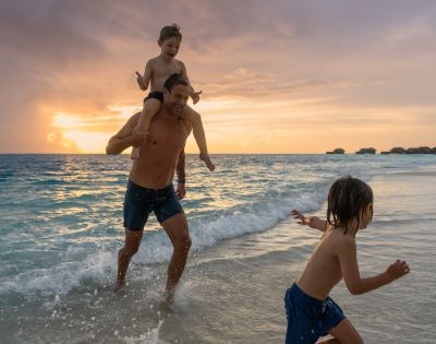 A joyful beach scene at sunset with a man carrying a child on his shoulders, running through gentle waves, while another child runs ahead, creating a playful and warm atmosphere.