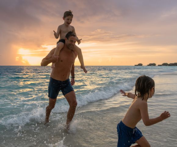 A joyful beach scene at sunset with a man carrying a child on his shoulders, running through gentle waves, while another child runs ahead, creating a playful and warm atmosphere.
