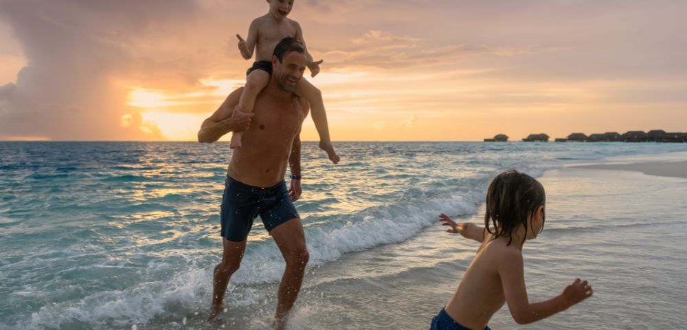 A joyful beach scene at sunset with a man carrying a child on his shoulders, running through gentle waves, while another child runs ahead, creating a playful and warm atmosphere.