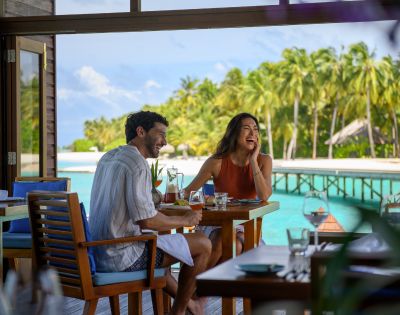 A couple enjoys a meal outdoors by a tropical beach. They are laughing, surrounded by lush palm trees and turquoise water, creating a joyful ambiance.