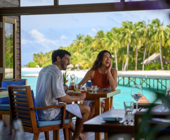 A couple enjoys a meal outdoors by a tropical beach. They are laughing, surrounded by lush palm trees and turquoise water, creating a joyful ambiance.