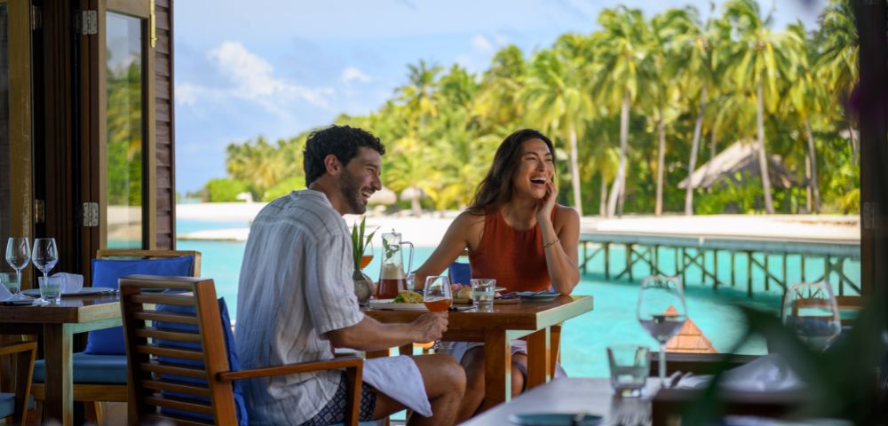 A couple enjoys a meal outdoors by a tropical beach. They are laughing, surrounded by lush palm trees and turquoise water, creating a joyful ambiance.