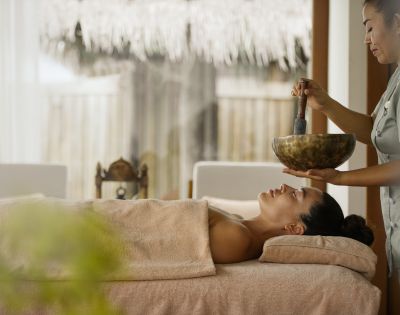 A person lies peacefully on a massage table, partially covered by a towel, while a therapist holds a Tibetan singing bowl, creating a serene spa atmosphere.