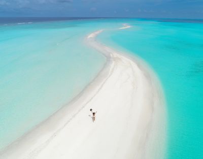 A person in swimwear is captured walking on a pristine, winding sandbar surrounded by turquoise waters under a clear blue sky, conveying a sense of serenity and solitude.