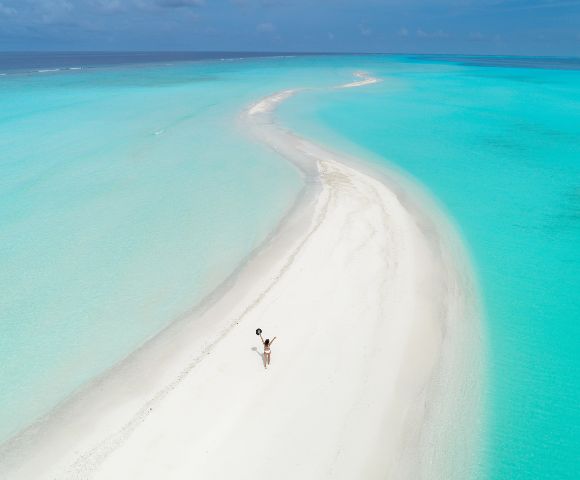 A person in swimwear is captured walking on a pristine, winding sandbar surrounded by turquoise waters under a clear blue sky, conveying a sense of serenity and solitude.