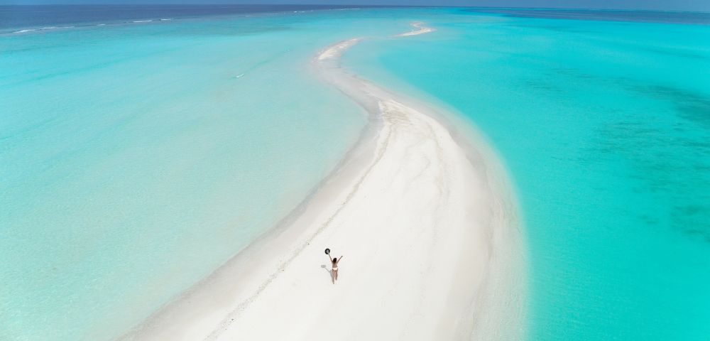 A person in swimwear is captured walking on a pristine, winding sandbar surrounded by turquoise waters under a clear blue sky, conveying a sense of serenity and solitude.