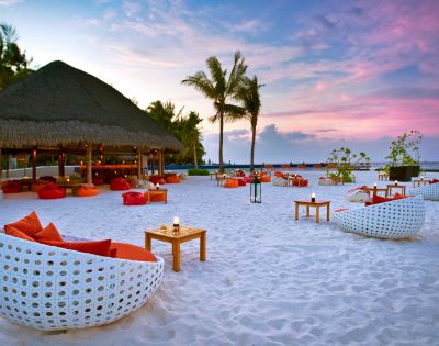A tropical beach at sunset, featuring white wicker chairs with orange cushions, wooden tables, palm trees, and a thatched-roof bar. Tranquil and inviting atmosphere.