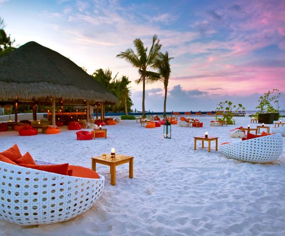 A tropical beach at sunset, featuring white wicker chairs with orange cushions, wooden tables, palm trees, and a thatched-roof bar. Tranquil and inviting atmosphere.
