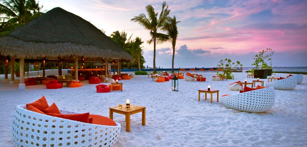 A tropical beach at sunset, featuring white wicker chairs with orange cushions, wooden tables, palm trees, and a thatched-roof bar. Tranquil and inviting atmosphere.
