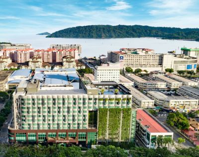 Arial view of a modern urban area with high-rise buildings, lush vertical gardens, and distant mountains by the sea under a clear blue sky.