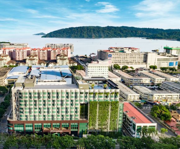Arial view of a modern urban area with high-rise buildings, lush vertical gardens, and distant mountains by the sea under a clear blue sky.