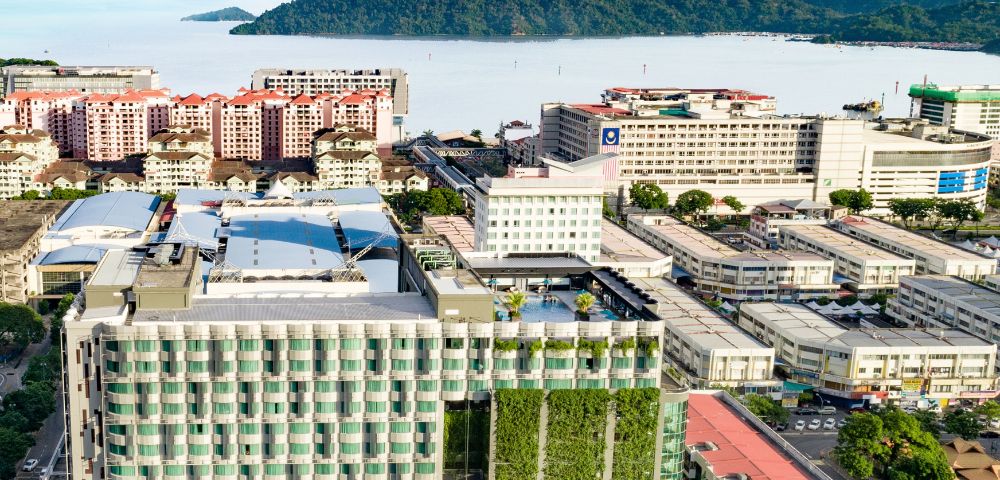 Arial view of a modern urban area with high-rise buildings, lush vertical gardens, and distant mountains by the sea under a clear blue sky.