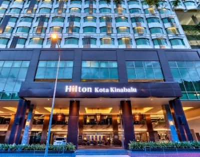 Modern hotel exterior with rows of windows and a warmly lit entrance. The sign reads 