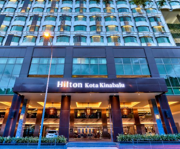 Modern hotel exterior with rows of windows and a warmly lit entrance. The sign reads 