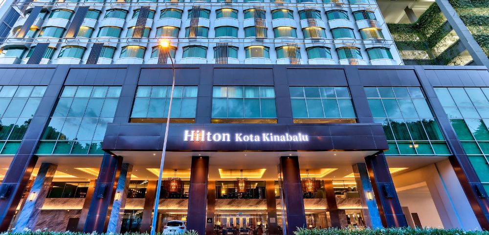 Modern hotel exterior with rows of windows and a warmly lit entrance. The sign reads 