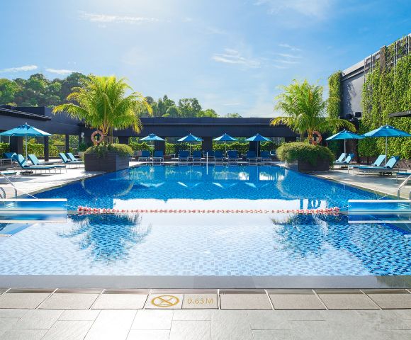 Luxurious outdoor pool with clear blue water, surrounded by lounge chairs and blue umbrellas. Palm trees and greenery create a tropical, relaxing atmosphere.