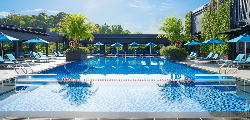 Luxurious outdoor pool with clear blue water, surrounded by lounge chairs and blue umbrellas. Palm trees and greenery create a tropical, relaxing atmosphere.