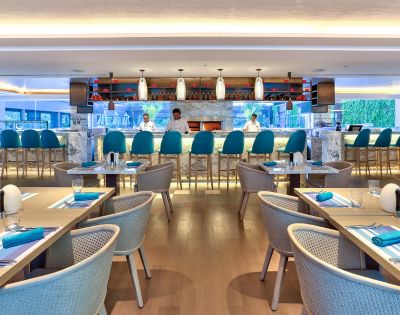 Modern restaurant interior with a sleek marble bar, teal chairs, and tables set for dining. Soft lighting creates a welcoming, elegant atmosphere.