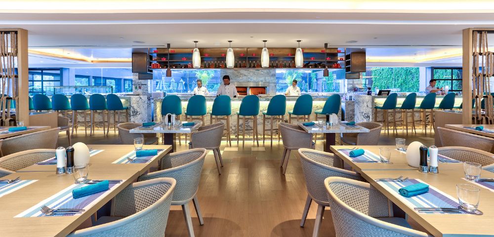 Modern restaurant interior with a sleek marble bar, teal chairs, and tables set for dining. Soft lighting creates a welcoming, elegant atmosphere.