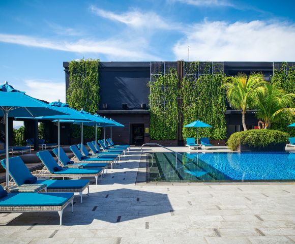 Lounge chairs with blue umbrellas line a poolside under a clear sky, alongside a building covered in lush green vines and palm trees, conveying a serene and tropical atmosphere.