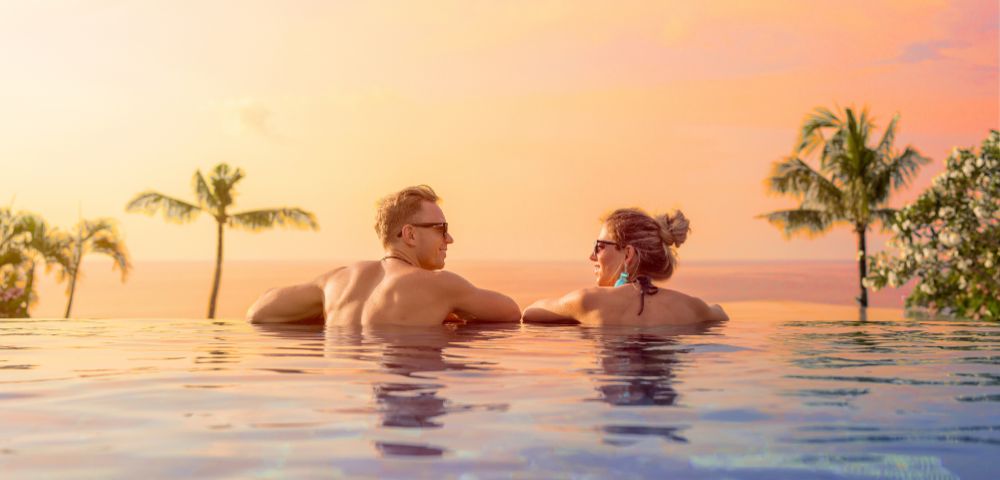 Two people relax in an infinity pool at sunset, gazing at each other. Palm trees and an orange-pink sky create a serene, tropical vibe.