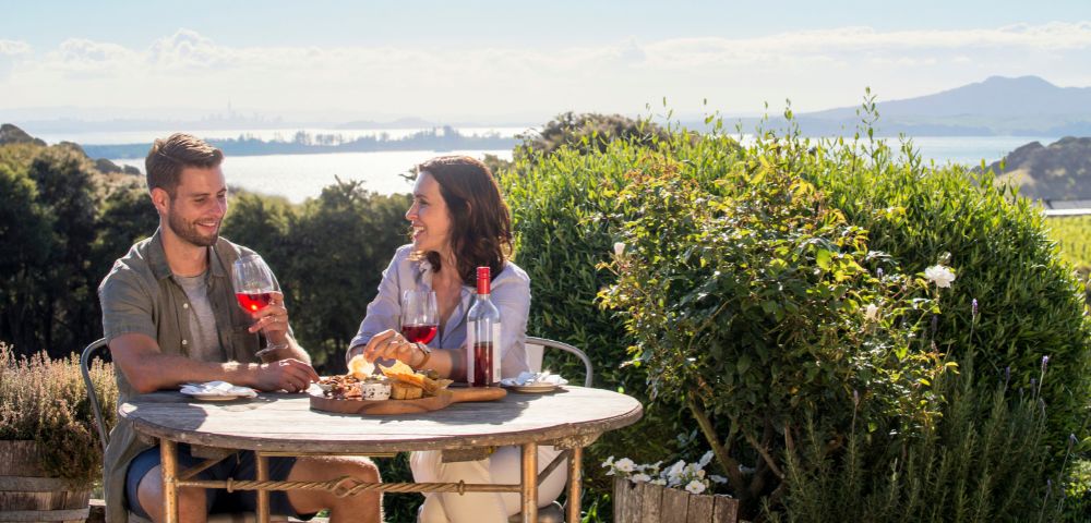 A couple enjoys wine and snacks at an outdoor table with a scenic view of lush greenery and distant mountains under a clear blue sky.