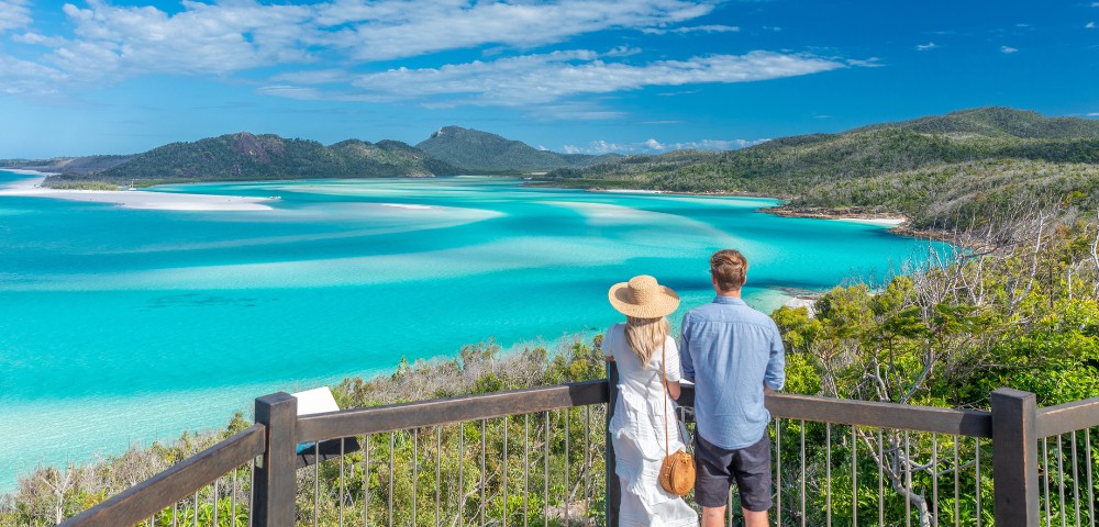 A couple stands on a wooden deck overlooking a breathtaking turquoise bay, surrounded by lush green hills under a clear blue sky. The scene is tranquil.
