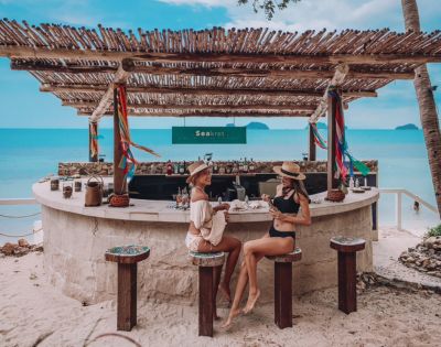 Two women in swimwear enjoy drinks at a rustic beach bar with a bamboo roof. They're surrounded by sand, colorful decor, and a serene ocean backdrop.