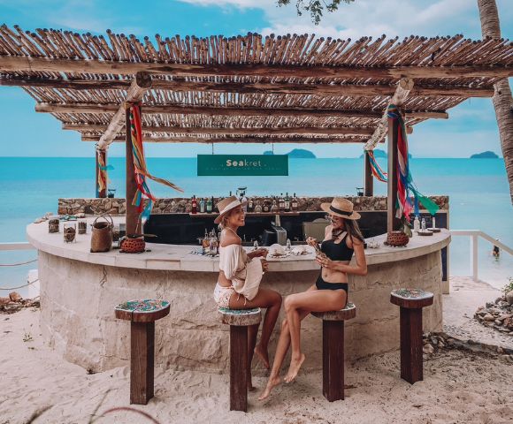 Two women in swimwear enjoy drinks at a rustic beach bar with a bamboo roof. They're surrounded by sand, colorful decor, and a serene ocean backdrop.