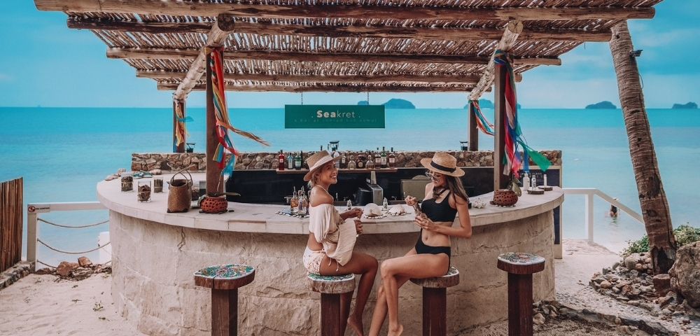 Two women in swimwear enjoy drinks at a rustic beach bar with a bamboo roof. They're surrounded by sand, colorful decor, and a serene ocean backdrop.