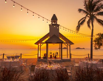 Sunset beach wedding scene with a decorated gazebo, string lights, and palm trees. Round tables with white tablecloths create a romantic atmosphere.