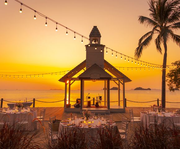 Sunset beach wedding scene with a decorated gazebo, string lights, and palm trees. Round tables with white tablecloths create a romantic atmosphere.