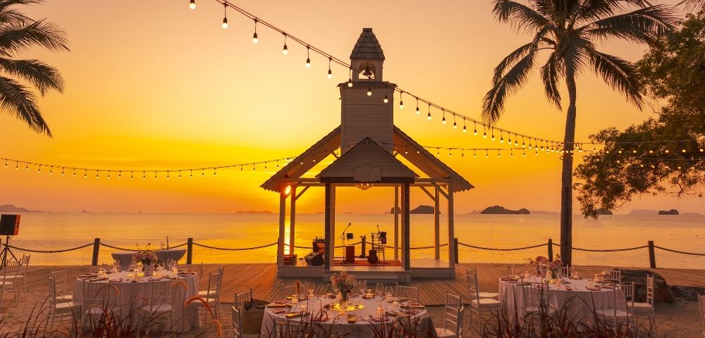 Sunset beach wedding scene with a decorated gazebo, string lights, and palm trees. Round tables with white tablecloths create a romantic atmosphere.
