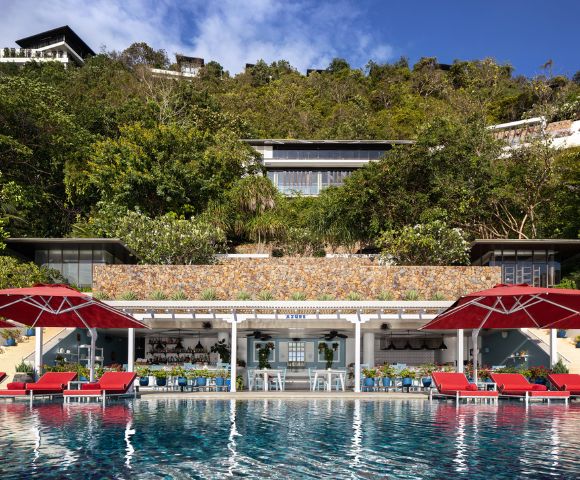 Luxurious poolside scene with red umbrellas and lounge chairs reflecting in a serene pool. Lush greenery and a chic modern building in the background.