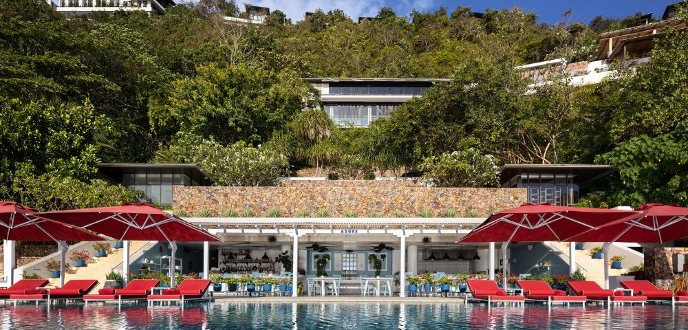Luxurious poolside scene with red umbrellas and lounge chairs reflecting in a serene pool. Lush greenery and a chic modern building in the background.