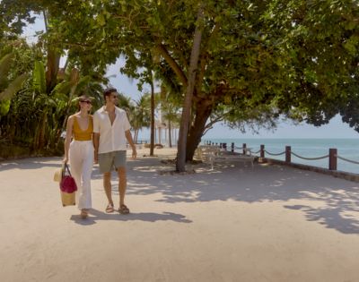 A couple strolls along a sunny beachside path lined with lush trees, carrying shopping bags. The scene conveys relaxation and leisure.