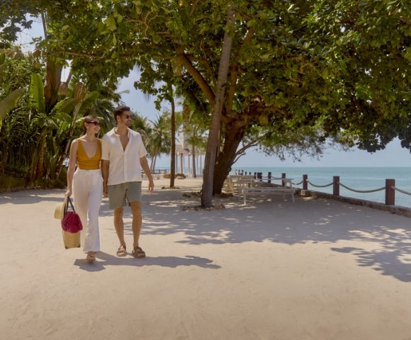 A couple strolls along a sunny beachside path lined with lush trees, carrying shopping bags. The scene conveys relaxation and leisure.