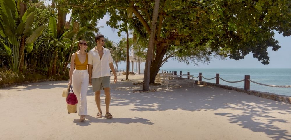 A couple strolls along a sunny beachside path lined with lush trees, carrying shopping bags. The scene conveys relaxation and leisure.
