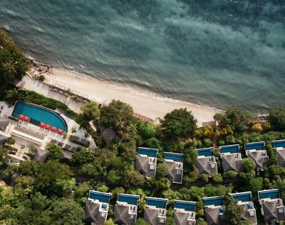 Aerial view of a tropical beachfront resort with villas and private pools surrounded by lush greenery. A serene, turquoise ocean contrasts the sandy shore.
