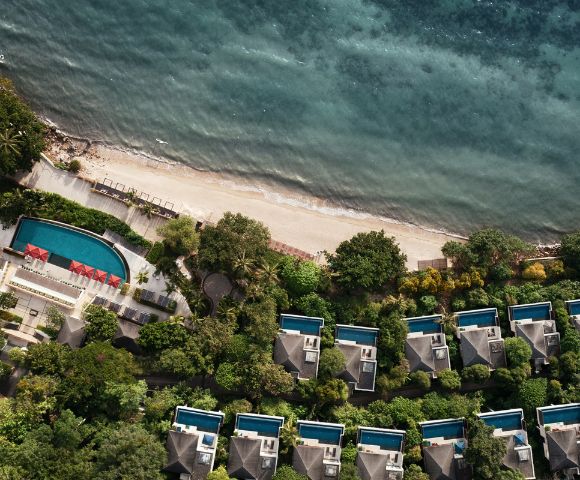 Aerial view of a tropical beachfront resort with villas and private pools surrounded by lush greenery. A serene, turquoise ocean contrasts the sandy shore.