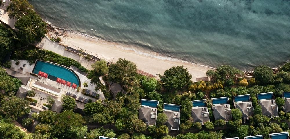 Aerial view of a tropical beachfront resort with villas and private pools surrounded by lush greenery. A serene, turquoise ocean contrasts the sandy shore.