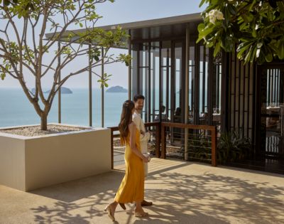 A couple walks on a sunny terrace overlooking the ocean. The woman wears a yellow dress, and the modern building nearby is partially covered by lush trees.