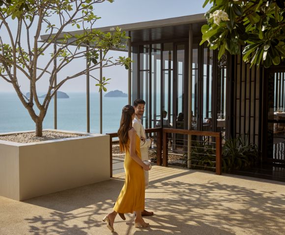 A couple walks on a sunny terrace overlooking the ocean. The woman wears a yellow dress, and the modern building nearby is partially covered by lush trees.