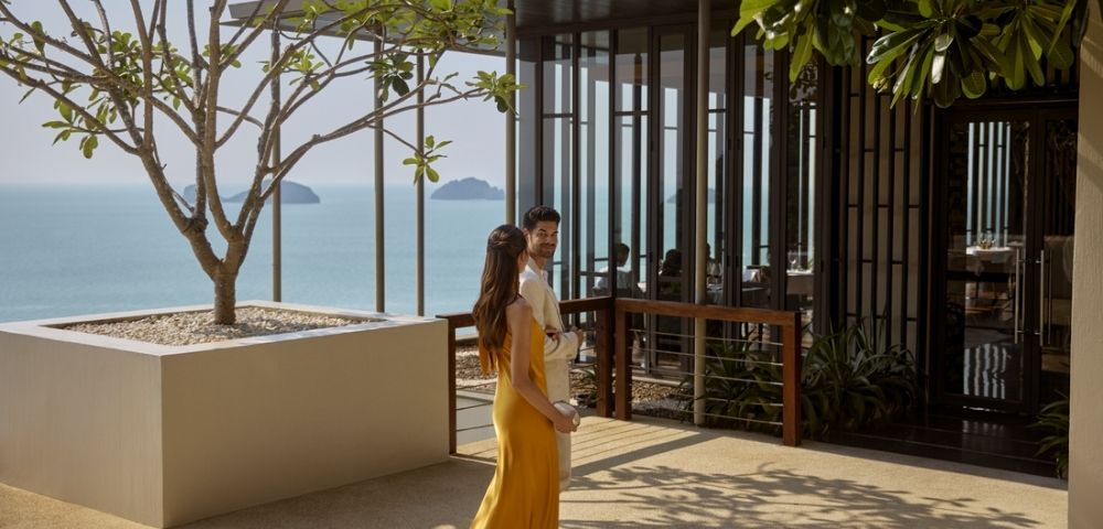 A couple walks on a sunny terrace overlooking the ocean. The woman wears a yellow dress, and the modern building nearby is partially covered by lush trees.