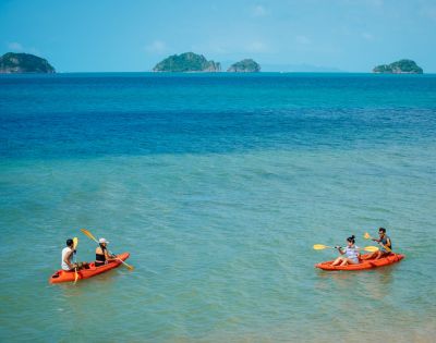 Two orange kayaks with people paddling on clear turquoise water, with small green islands in the distance under a sunny blue sky.